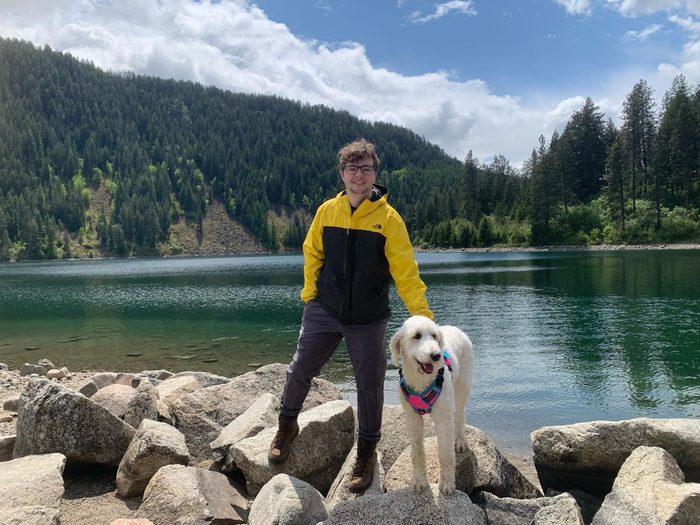 Delaney Mizell standing on boulders at the edge of a mountain lake in a yellow and black jacket, one hand resting on a white curly-haired dog beside him, with a pine-covered slope and broken cloud behind.