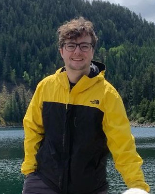 Delaney Mizell in glasses and a yellow and black rain jacket, smiling at the camera at the edge of a mountain lake, with a pine-covered slope behind him and the white coat of his dog just in frame at his side.