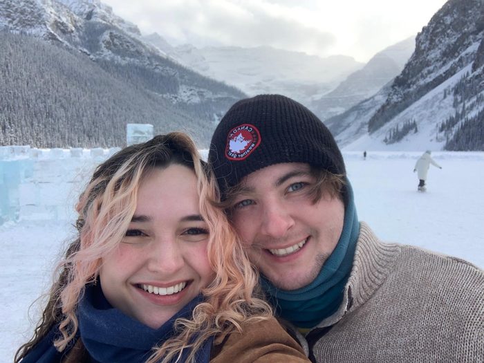 Delaney Mizell and his wife Devon, cheek to cheek and smiling into the camera, standing on a frozen lake ringed by snow-covered peaks and dark evergreens. He is in a dark knit cap, she is in a blue scarf, and a skater is out on the ice behind them.
